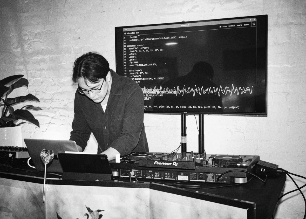 Brandon Tai livecoding at a laptop and DJ controller, a Strudel patch on the screen behind him.